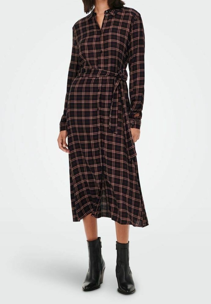 Eine Frau trägt ein schwarzes und rotes kariertes langärmliges Midikleid mit einem Taillengürtel und schwarzen Stiefeln bis zur Knöchel, während sie vor einem weißen Hintergrund steht.