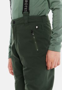 Zielone spodnie z zamkiem błyskawicznym i regulowanymi szelkami, o smukłym kroju i gładkiej fakturze. Mały detal z logo na przedniej kieszeni.