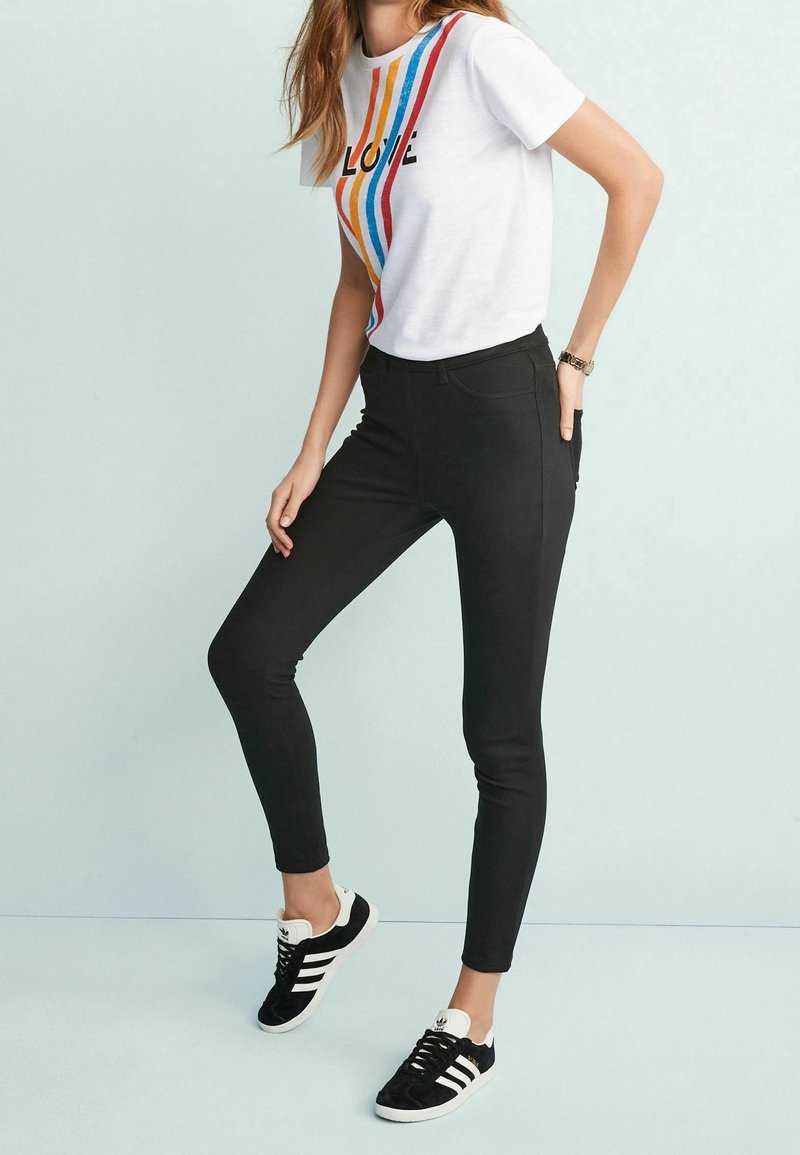 Femme debout portant un t-shirt blanc à rayures colorées avec le texte "LOVE", pantalon slim noir et baskets Adidas noires et blanches.