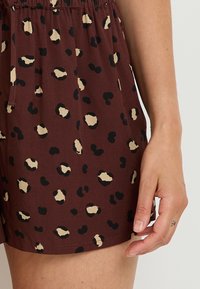 Shorts en tissu brun avec des taches de léopard noires et beiges, dotés d'une taille élastique et de poches latérales. Texture lisse, design décontracté.