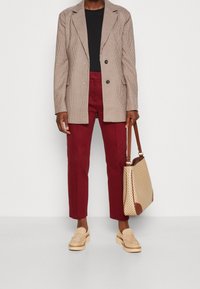 Blazer de cuadros beige, camiseta negra, pantalones burdeos a medida, mocasines marrones, llevando un bolso de mano de patrón claro con acentos marrones.