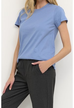 Kvinde iført en lyseblå t-shirt med korte ærmer og mørkegrå bukser, stående med den ene hånd i lommen mod en ensfarvet baggrund.