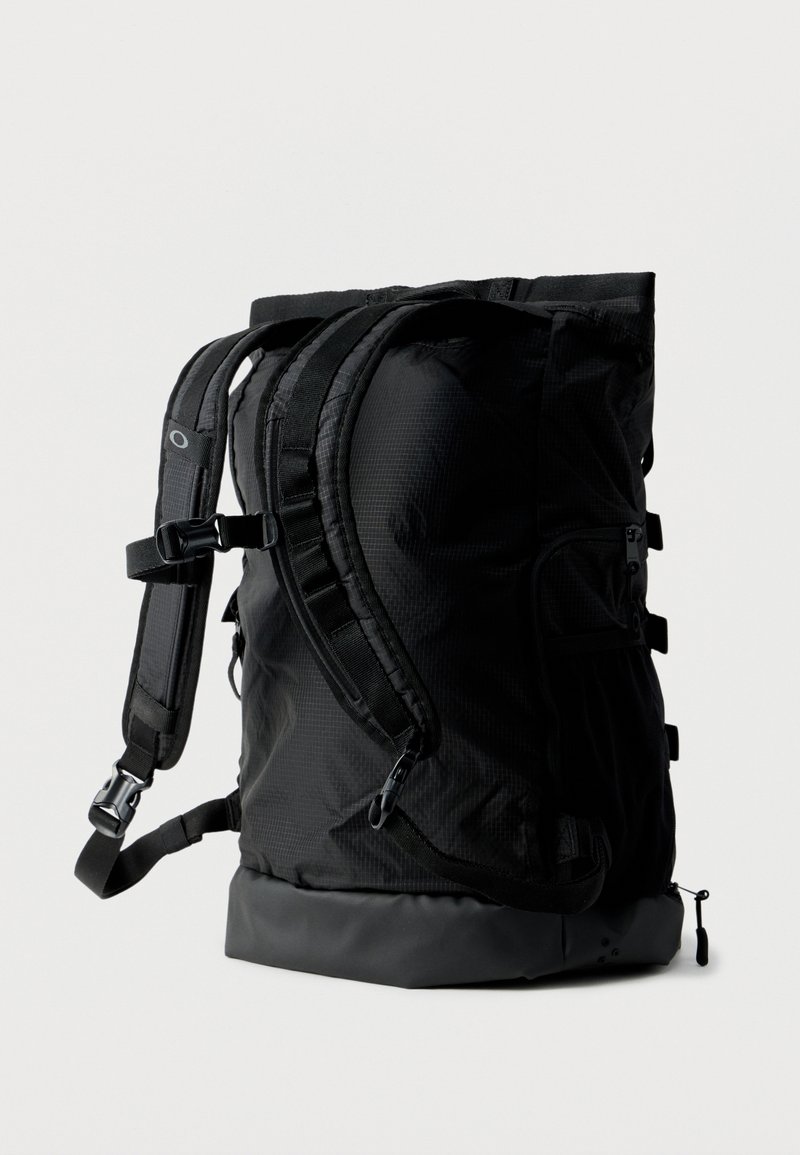 Sac à dos noir à fermeture roulante avec bretelles rembourrées, multiples boucles et compartiments zippés, debout sur un fond blanc.