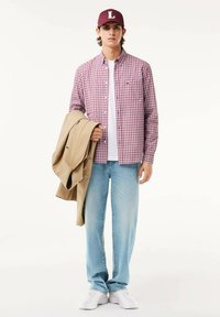 Camisa de cuadros en rojo y blanco, superpuesta sobre una camiseta blanca. Jeans de color azul claro y zapatos blancos tipo slip-on. Chaqueta beige colgada del brazo.