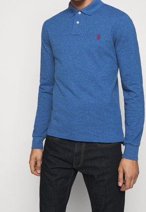 Mann trägt ein blaues langärmeliges Polohemd mit kleinem roten gesticktem Logo auf der Brust und dunkle Jeans.