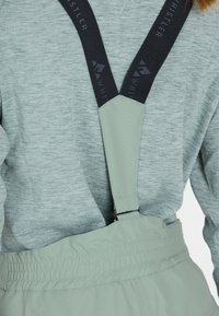 Hellgrauer Pullover unter mintgrünen Latzhosen mit schwarzen verstellbaren Trägern; die Träger sind mit Branding versehen und haben eine glatte Textur.