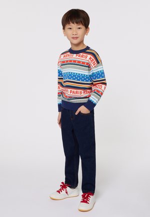 Pull en tricot coloré avec des motifs bleus, rouges et verts ; jeans en denim ; baskets blanches avec des accents rouges ; posture debout.