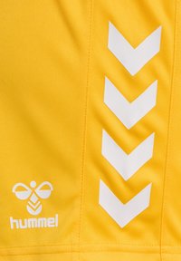Camiseta deportiva amarilla con una textura suave. Presenta patrones de chevron blancos y un logotipo en la esquina inferior izquierda.