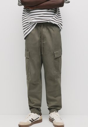 Pantaloni cargo verzi măslinii cu talie elastică și tiv, dotati cu mai multe buzunare laterale, fabricați dintr-un material durabil. Asociați cu adidași bej.