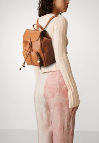 Sac à dos en cuir marron avec un rabat repliable, fermeture à cordon, et poche zippée à l'avant. Il présente des bords arrondis et des accents matériels minimalistes.