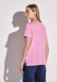 Pink cotton t-shirt with a relaxed fit, short sleeves, and side slits. Model shown from the back, wearing dark blue jeans.