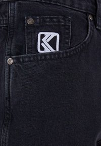 Czarne jeansy z denimu z naszywaną kieszenią ozdobioną białym haftowanym logo, srebrnymi elementami ozdobnymi oraz teksturowanym wykończeniem tkaniny.