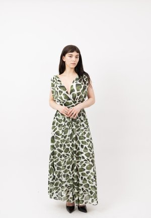 Mujer con cabello largo y oscuro que lleva un vestido maxi sin mangas con estampado verde y blanco y tacones negros puntiagudos, de pie sobre un fondo blanco.