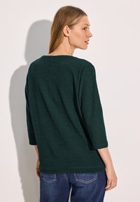 Haut texturé vert foncé à manches trois-quarts, col rond et coupe ample ; associé à un jean en denim bleu.
