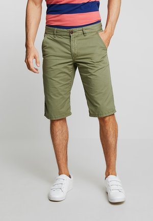 Olivgrüne Baumwollshorts mit mehreren Taschen, einem Knopfverschluss und gesäumten Kanten. Tragen mit weißen Turnschuhen und gestreiftem Shirt.