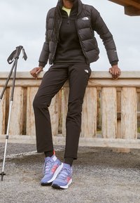 Doudoune noire matelassée, couche intermédiaire noire zippée, pantalon noir et chaussures de sport grises avec des accents rouges. Des bâtons de marche reposent contre une rambarde en bois.