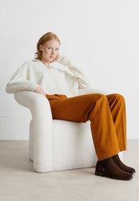 Crème trui, roestkleurige, wijdvallende broek en bruine enkellaarsjes. Model zit in een witte, gestructureerde fauteuil tegen een neutrale achtergrond.