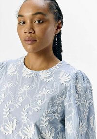 Top de tela azul claro con bordado floral blanco, cuello redondo, mangas abullonadas y un detallado de costura intrincada en todo.
