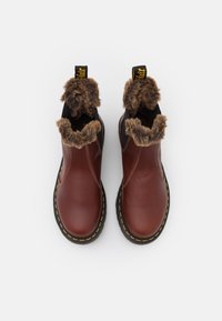 Botas de tobillo de cuero marrón con forro de piel sintética, lados elásticos negros y costuras amarillas a lo largo de la suela. Cuentan con lengüetas para facilitar su uso.