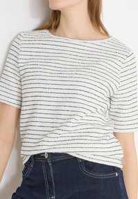 Femme portant un haut en maille texturée blanc à manches courtes avec de fines rayures horizontales noires et un jean denim bleu foncé.