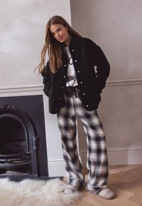 Veste en polaire noire avec des boutons blancs, portée sur un t-shirt blanc à motif graphique. Pantalon large à carreaux beige et noir, assorti à des chaussons beiges.