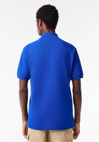 Polo bleu avec col, manches courtes et tissu texturé. Coupe décontractée, présenté de dos sur un fond neutre.