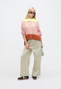 Megzta rožinės ir oranžinės spalvų megztinį su sagotu apykakle, derinama su plačiomis žaliomis languotomis kelnėmis ir juodais sportbačiais su rudos spalvos padais.