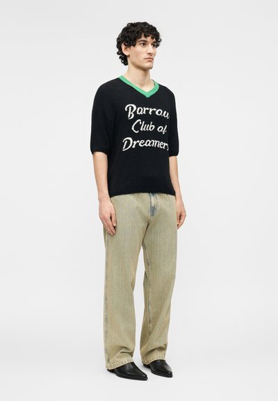 Νέος άνδρας με σγουρά μαλλιά που φοράει μαύρο πουλόβερ με κοντά μανίκια, πράσινο γιακά και κείμενο "Barrow Club of Dreamers", μπεζ παντελόνι και μαύρα παπούτσια.
