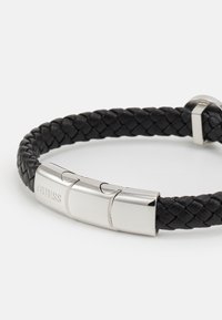 Svart flettet lærarmbånd med en skinnende sølvfarget lås. Har gravert "GUESS"-logo på metalldelen.