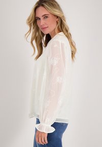 Witte sheer blouse met bloemenborduursel, lange puffmouwen en gerimpelde manchetten. Gecombineerd met blauwe jeans voor een casual look.