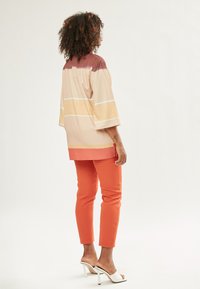 Blusa colorida y de gran tamaño con rayas horizontales en marrón, beige y amarillo. Combinada con pantalones ajustados naranjas y sandalias de tacón blancas.