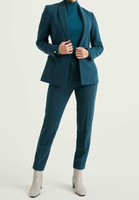 Teal skräddarsydd kavaj och matchande byxor i ett strukturerat tyg; figursydd design; kombinerad med beige ankelskor. Snittet ger en modern look.