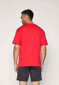 Mand vendt v�æk, iført en ensfarvet rød t-shirt med korte ærmer og sorte shorts mod en neutral baggrund.
