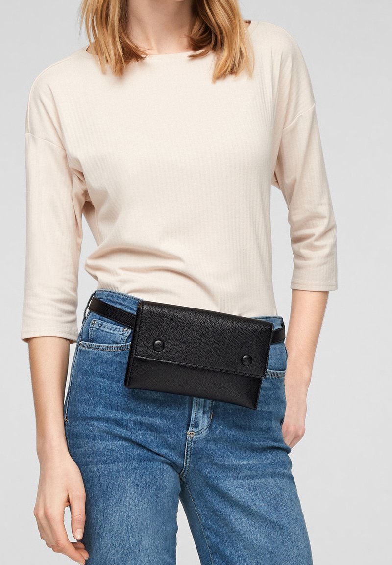 Sac banane en cuir noir avec fermetures à boutons, forme rectangulaire plate, porté sur un haut beige clair et un jean bleu. Texture lisse, design minimaliste.