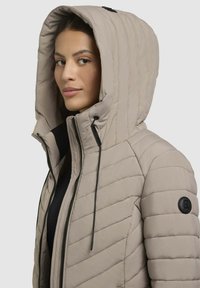 Chaqueta aislante beige con un diseño acolchado, cuello alto, cierre de cremallera en la parte delantera y capucha con cordón. Presenta un parche de logo negro en la manga.