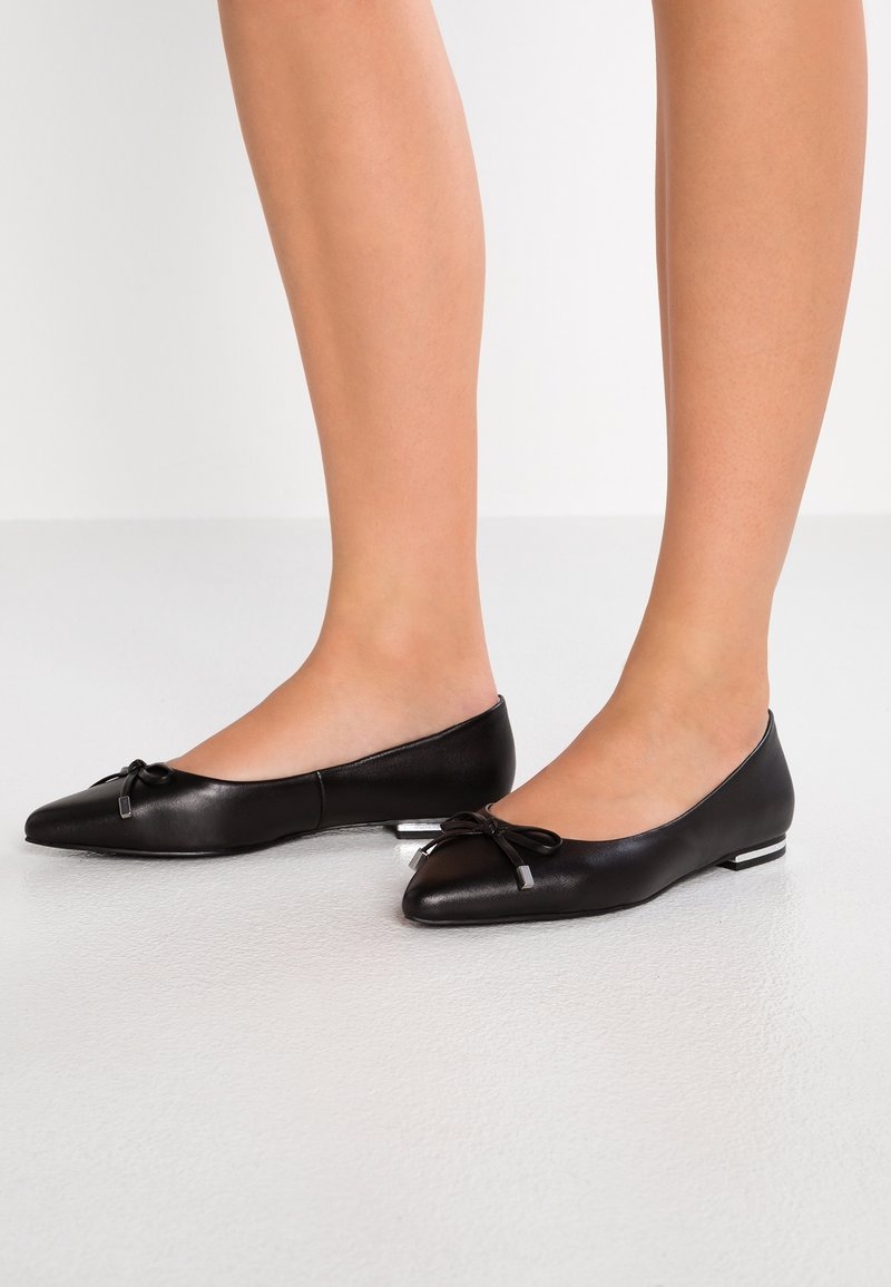 Ballerines en cuir noir avec un bout arrondi, ornées d'un nœud à l'avant et d'un petit talon, présentant une texture lisse et un design minimaliste.