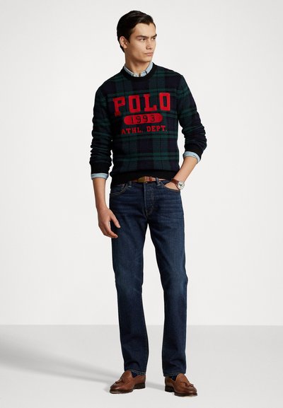 Suéter a cuadros en verde oscuro y azul marino con texto rojo "POLO", combinado con jeans azules y mocasines marrones. El modelo se encuentra de pie con una mano en la cadera.