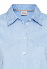 Chemise bleu clair à boutons avec col pointu et poche poitrine visible, avec une petite étiquette blanche à l'intérieur de l'encolure.