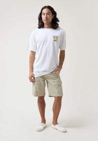 Camiseta de algodón blanca con un gráfico amarillo y azul en el pecho, combinada con pantalones cortos tipo cargo beige y zapatillas de lona blancas.