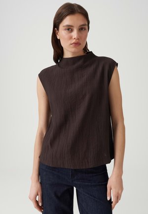 Jeune femme aux cheveux bruns lisses portant un haut texturé sans manches marron foncé et un jean bleu foncé devant un fond uni.