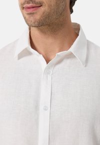 Weißes Leinenhemd mit klassischem Kragen, ausgestattet mit einer durchgehenden Knopfleiste und einem strukturierten Stoff. Die Knöpfe sind transparent und verleihen einen dezenten Akzent.