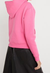 Sudadera con capucha de forro polar en un rosa brillante, con puños y dobladillo acanalados. Combinada con una falda de cuero negro con bolsillo trasero visible.