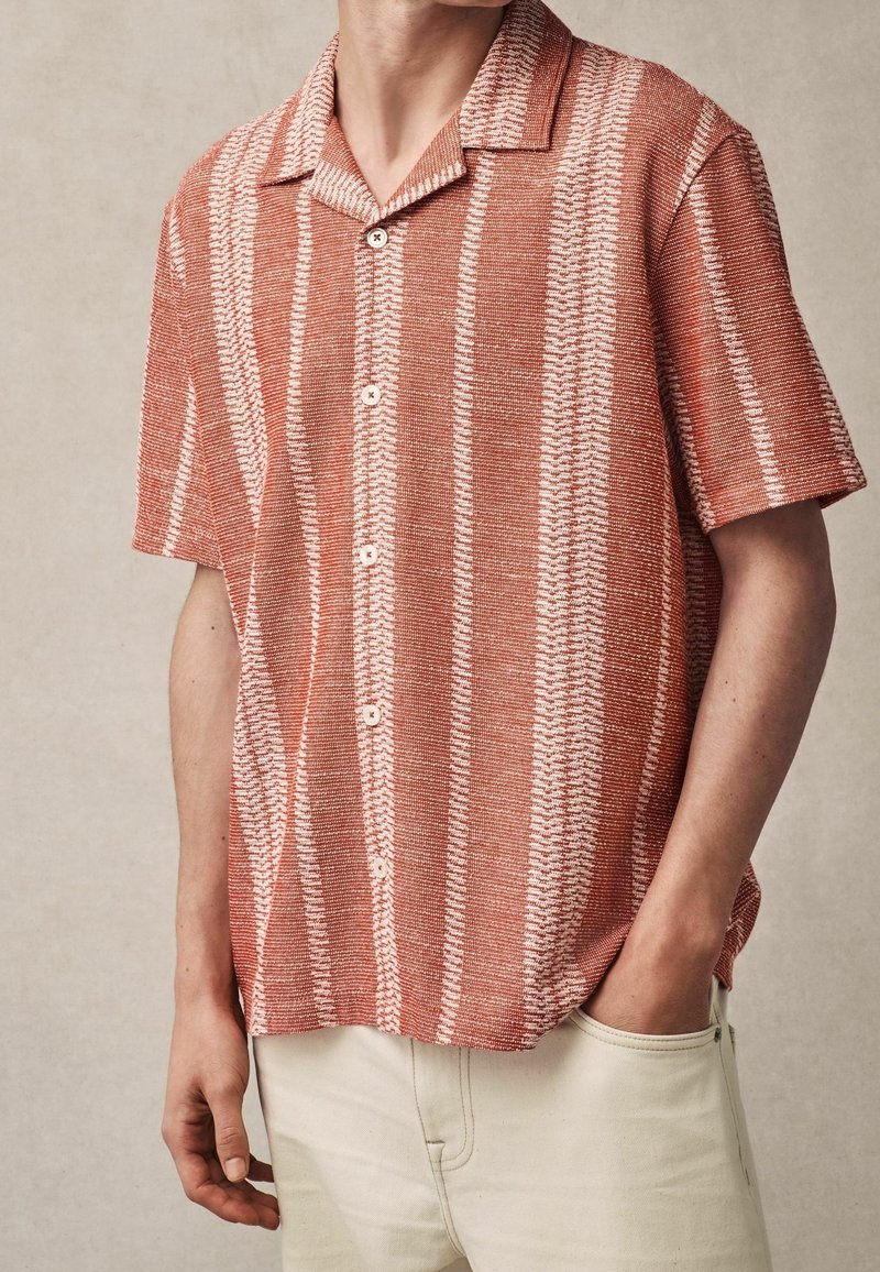 Homme portant une chemise à manches courtes boutonnée avec des rayures verticales rouges et blanches, associée à un pantalon clair, une main dans la poche.