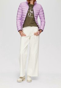 Donna che indossa una giacca a vento leggera di colore viola chiaro sopra una maglietta grafica oliva, pantaloni bianchi a gamba larga e stivaletti di pizzo color crema, in piedi con le mani sui fianchi.