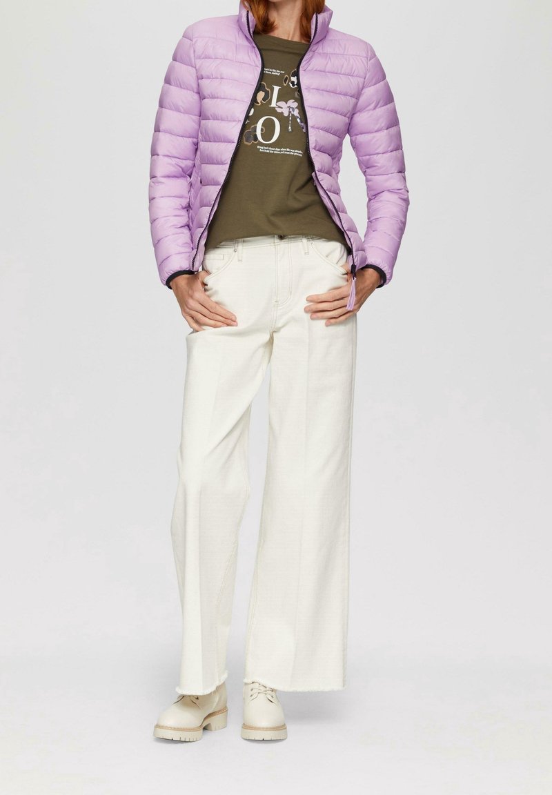 Donna che indossa una giacca a vento leggera di colore viola chiaro sopra una maglietta grafica oliva, pantaloni bianchi a gamba larga e stivaletti di pizzo color crema, in piedi con le mani sui fianchi.