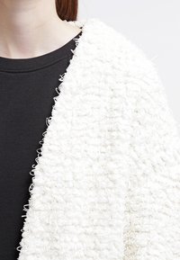 Bílý texturovaný kardigan z měkké smyčkové tkaniny. Nosený přes černý top, s volným střihem a vrstveným designem.