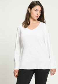 Bílá halenka s dlouhým rukávem ve tvaru V vyrobená z měkké látky, s uvolněným střihem a hladkou texturou, zkombinovaná s černými kalhotami.