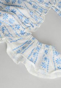 Tessuto bianco con motivi floreali blu e sottili strisce verticali. I bordi arricciati e le aree raccolte aggiungono texture e dettagli.