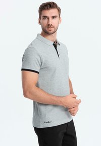 Šedé polo tričko s krátkým rukávem, černým límcem a manžetami, s knoflíkovou légou a vyšívaným logem blízko dolního okraje. Měkký bavlněný materiál.