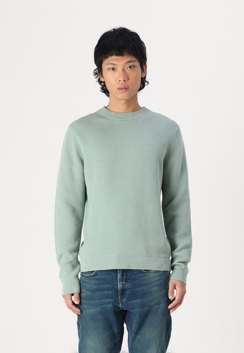 Suéter de malha verde-clara com decote redondo e punhos canelados, usado com jeans de ganga azul. A textura parece suave e respirável.